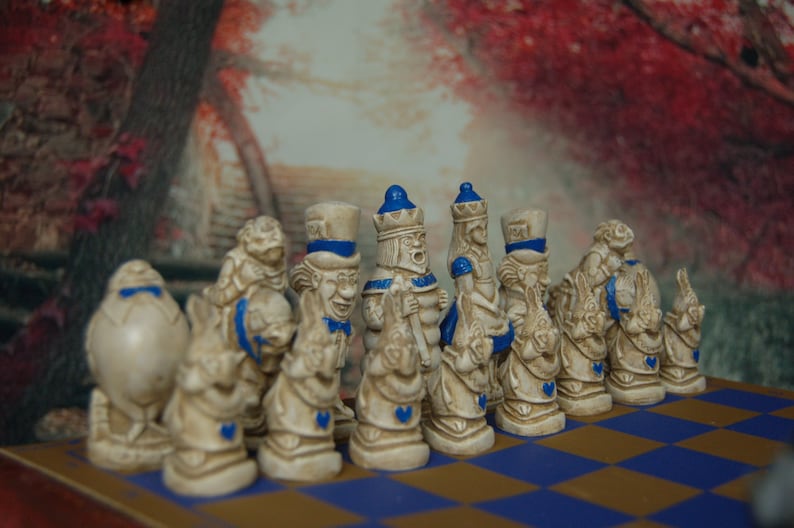 May include: A set of white ceramic chess pieces with blue accents, depicting characters from Alice in Wonderland. The pieces are arranged on a blue and brown checkered board.