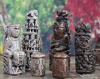 May include: A collection of intricately carved chess pieces in various shades of gray and brown. The pieces depict figures such as a queen, king, and rooks, with detailed carvings and a weathered appearance. The chess set is displayed against a blurred background.
