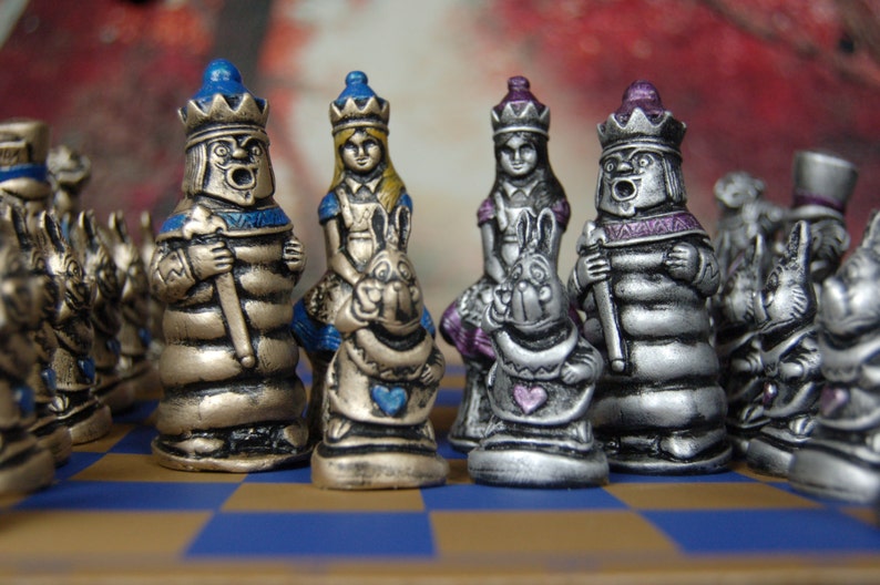 May include: A silver and gold chess set featuring characters from Alice in Wonderland. The pieces include the Queen of Hearts, the Mad Hatter, and the White Rabbit. The chessboard is a black and white checkered pattern.