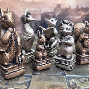 Puede incluir: Un conjunto de siete figuras de animales, incluyendo un conejo, un pato, un zorro, un gato y una ardilla, cada uno sobre una pila de libros. Las piezas de ajedrez tienen un acabado en bronce y plata, dispuestas sobre un tablero.