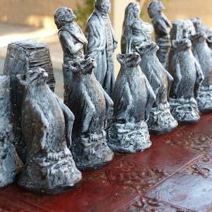 May include: A unique chess set featuring hand-painted, gray and white chess pieces. The pieces are sculpted in a fantasy theme, with figures and creatures. The set is displayed on a red, patterned chessboard, creating a striking contrast.