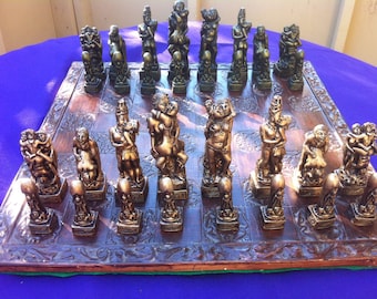 May include: A detailed chess set with bronze-colored pieces depicting various human figures in intimate poses. The chess board is made of dark wood with carved edges, set against a blue background.