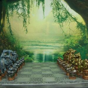 May include: A fantasy-themed chess set with detailed, sculpted pieces in shades of gray and gold. The chess board is gray with a patterned design. The background features a forest scene.