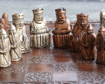 Puede incluir: Un juego de ajedrez vintage con piezas de color plateado y cobre sobre un tablero de madera con un diseño intrincado. Las piezas tienen forma de figuras medievales.