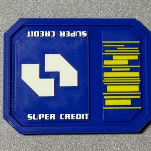 May include: A blue rectangular badge with the words "SUPER CREDIT" in white. The badge has a white geometric design and yellow horizontal lines. The badge is on a gray surface.