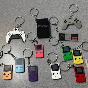 May include: A collection of 17 colorful keychains featuring various video game controllers and handheld game consoles. The keychains are made of plastic and feature a variety of colors, including red, blue, green, yellow, and white. The keychains are all attached to silver key rings.