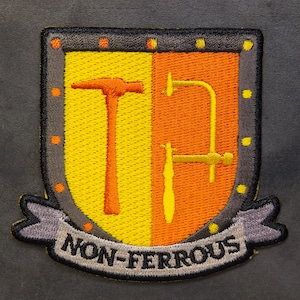 May include: An embroidered patch in grey and black, featuring a yellow and orange shield. The shield depicts a hammer and a pair of pliers. The text "NON-FERROUS" is displayed on a grey banner below the shield.
