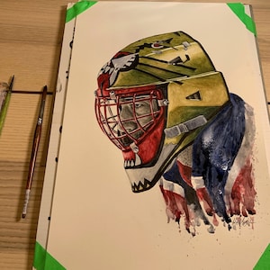 May include: Watercolor painting of a hockey goalie's mask with a green, yellow, and red color scheme. The mask has a fierce, animalistic design with sharp teeth and a menacing expression. The background is a blurred watercolor wash of blue and white, suggesting a hockey jersey.