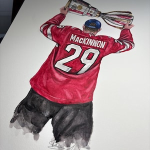 May include: Watercolor painting of a hockey player wearing a red jersey with the number 29 and the name "MacKinnon" on the back. The player is holding a trophy above their head.