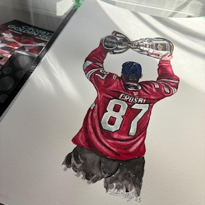May include: Watercolor painting of a hockey player in a red jersey with the number 87 and the name "CROSBY" on the back, holding a silver trophy above his head. The player is viewed from the back, wearing a blue cap. The artwork is on white paper.