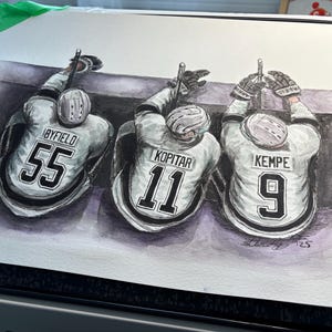 Può includere: Dipinto ad acquerello di tre giocatori di hockey nelle loro maglie. I giocatori indossano maglie con i numeri 55, 11 e 19. I nomi dei giocatori sono Byfield, Kopitar e Kempe.