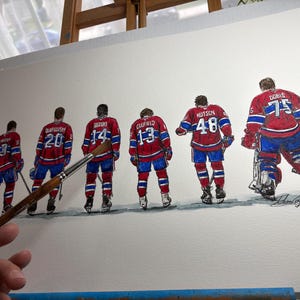 Puede incluir: Pintura a la acuarela de seis jugadores de hockey sobre hielo con uniformes rojos, blancos y azules, vistos por la espalda. Cada camiseta muestra un n&uacute;mero diferente. Un pincel se sostiene frente a la obra de arte. La pintura est&aacute; sobre un lienzo blanco.