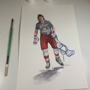 May include: A watercolour painting of a hockey player wearing a grey jersey with the text "DETROIT Red Wings" and red trousers. The player is holding a hockey stick and a pair of ice skates. The painting is signed "Hourdequin '21".