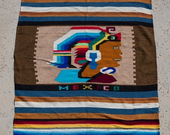 May include: A rectangular woven textile featuring horizontal stripes in shades of blue, brown, white, red, and yellow. The centre displays a profile illustration of a person wearing a colourful headdress, with the word MEXICO printed below.