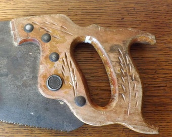 Könnte beinhalten: Eine antike Handsäge mit einem Holzgriff. Der Griff hat ein geschnitztes Weizenmuster und Metallakzente. Die Klinge ist aus Stahl und hat eine leichte Krümmung.
