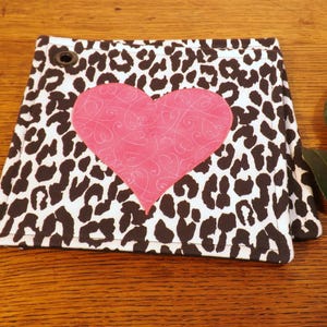 May include: A black and white leopard print fabric hot pad with a pink heart in the center. The heart has a swirl design.