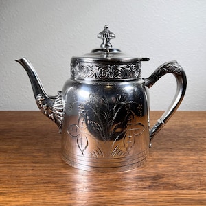 May include: A silver teapot with an ornate design and a floral pattern. The teapot has a lid with a knob on top and a spout on the side. The handle is curved and has a decorative design.