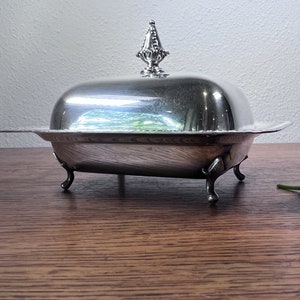 May include: A silver butter dish with a domed lid and a decorative finial. The dish has four feet and is sitting on a wooden surface.
