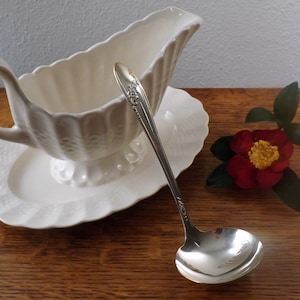 May include: A white ceramic gravy boat with a matching saucer and a silver ladle. The gravy boat has a fluted design and a handle. The ladle has a decorative handle.