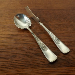 May include: A set of two silver-plated sugar serving utensils. The spoon has a decorative, scalloped bowl and a twisted handle. The knife has a decorative, floral design on the handle.
