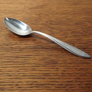 May include: A silver teaspoon with a rounded bowl and a simple, elegant design. The spoon has a slightly curved handle with a decorative pattern.
