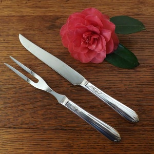 May include: A silver carving set with a knife and fork on a wooden surface. The set has a decorative design on the handles.