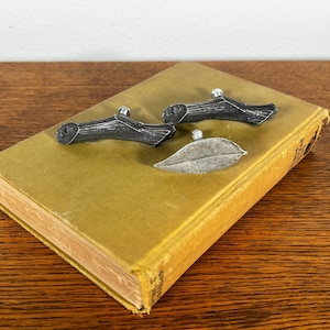 May include: Three metal drawer pulls in the shape of a twig and a leaf. The pulls are silver in color and are arranged on a yellow book.