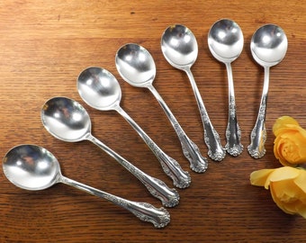 May include: Seven silver spoons with ornate handles arranged in a fan shape on a wooden surface. The spoons are polished and reflect light. Two yellow roses are in the background.