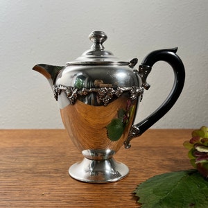 May include: Silver creamer with a black handle and grapevine design. The creamer has a round body and a pedestal base.