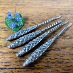 May include: Four silver, decorative,  nail cleaning tools with a textured, diamond-shaped pattern. The tools are arranged in a row on a wooden surface.