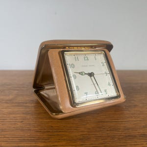 May include: A vintage tan leather-covered travel alarm clock with a white face and gold-colored hands. The clock face reads "Premier - Made in USA" and "Electric".