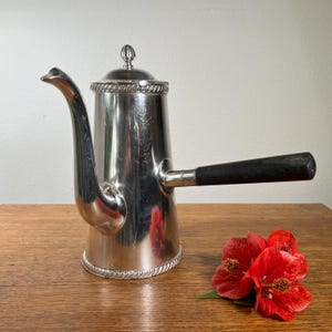 May include: A silver-colored coffee pot with a long, curved spout and a black handle. The pot has a decorative rim and a small, ornate lid. A cluster of red flowers rests on the wooden surface next to the pot.