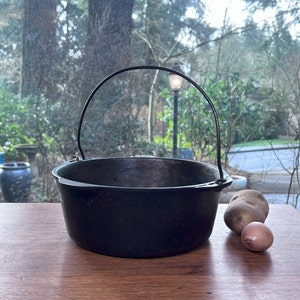 May include: A black cast iron pot with a single handle. The pot is sitting on a wooden surface with two potatoes and an onion beside it.