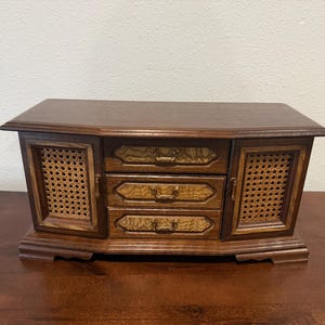 May include: A vintage wooden jewelry box with a curved top and three drawers. The box features woven cane panels on the sides and decorative brass hardware. The wood has a rich, warm brown finish.