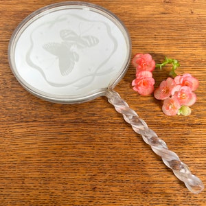 May include: A vintage hand mirror with a clear glass handle and a round, etched glass mirror. The mirror has a delicate butterfly design etched into the glass. The mirror is lying on a wooden surface with pink flowers.