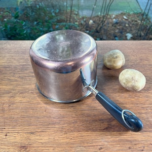 May include: A silver saucepan with a black handle and a copper-colored interior. The saucepan is sitting on a wooden surface with two potatoes next to it.