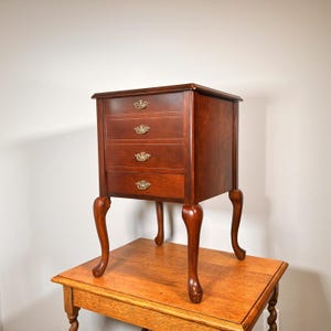 May include: A small, antique wooden chest of drawers with four drawers and brass hardware. The chest is a rich, dark brown color and has curved legs. It is sitting on top of a wooden table.
