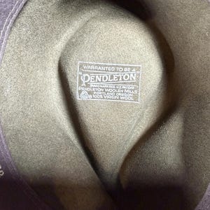 May include: Close-up of the interior of a green wool hat. The hat features a label that reads "WARRANTED TO BE A PENDLETON" and "100% VIRGIN WOOL". The hat's brim is a darker shade of green. The hat's style is a fedora.