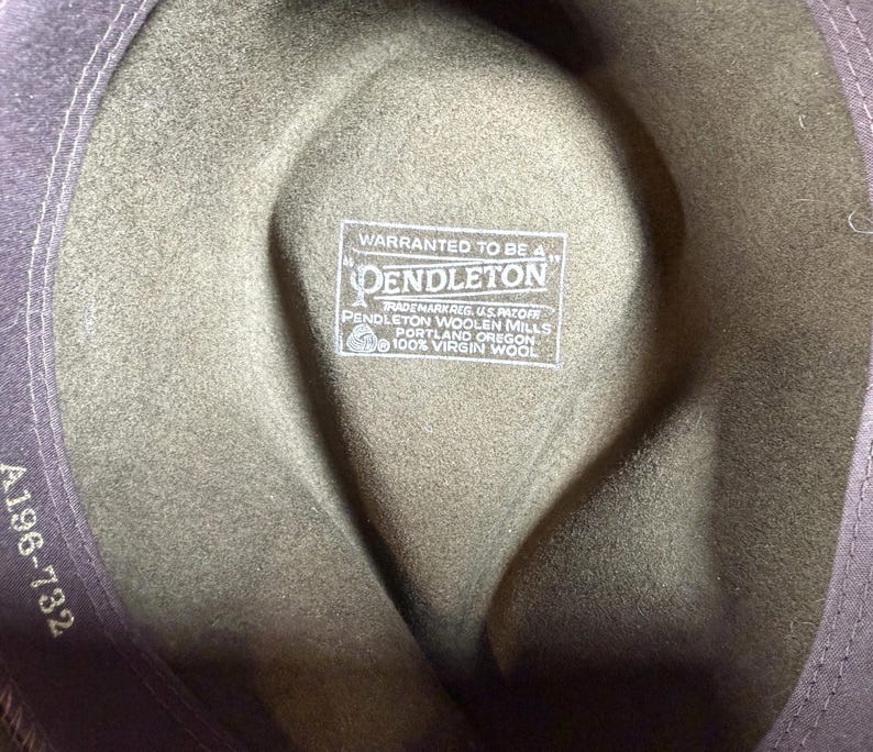 May include: Close-up of the interior of a green wool hat. The hat features a label that reads "WARRANTED TO BE A PENDLETON" and "100% VIRGIN WOOL". The hat's brim is a darker shade of green. The hat's style is a fedora.