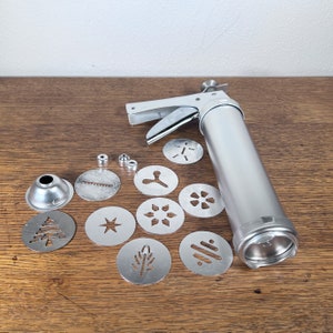 May include: A silver cookie press with a variety of metal cookie cutters, including a star, snowflake, tree, and leaf. The cookie press has a lever and a barrel for filling with cookie dough.