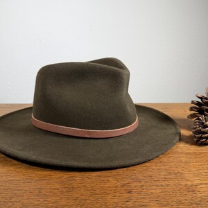 May include: A dark olive green fedora hat with a brown leather band. The hat has a wide brim and a classic, structured crown. A pine cone sits to the right of the hat on a wooden surface.