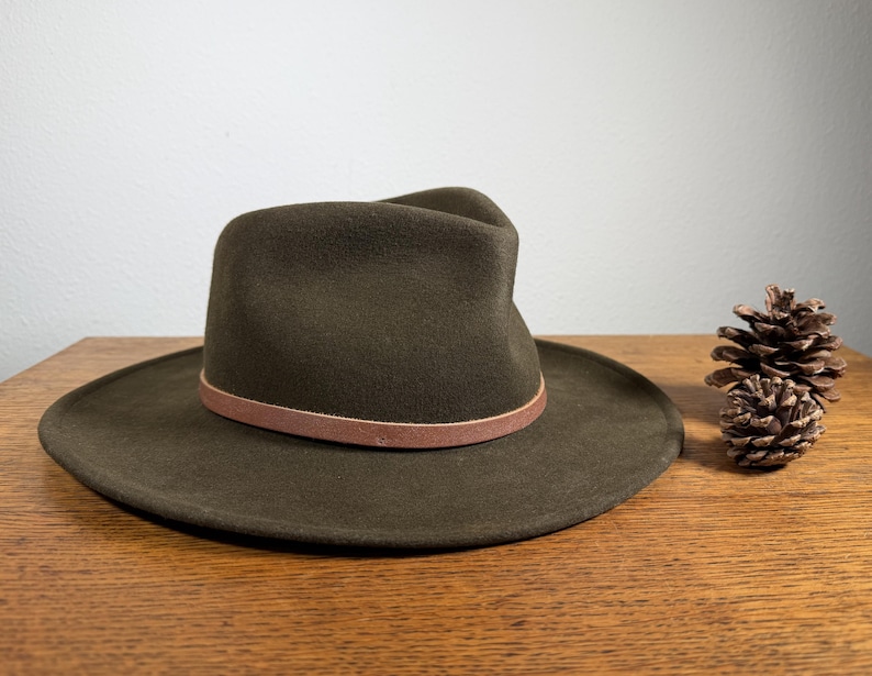 May include: A dark olive green fedora hat with a brown leather band. The hat has a wide brim and a classic, structured crown. A pine cone sits to the right of the hat on a wooden surface.