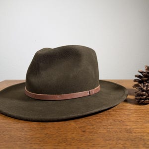 May include: A dark olive green fedora hat with a brown leather band. The hat sits on a wooden surface next to a pine cone. The hat has a wide brim and a rounded crown.