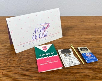 May include: A collection of sewing supplies, including Singer hand sewing needles, a card that reads "A Gift of Gold", and other sewing needles. The items are arranged on a wooden surface.