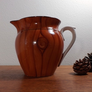 May include: A brown ceramic pitcher with a woodgrain pattern and a white handle. The pitcher is sitting on a wooden surface.