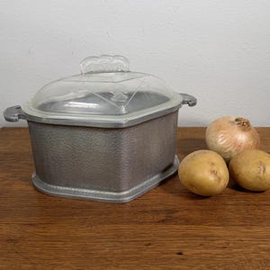 May include: A rectangular aluminum cooking pot with a textured exterior finish and two side handles. The pot includes a clear, molded glass lid featuring a decorative embossed pattern on the top surface. The pot is positioned next to two potatoes.