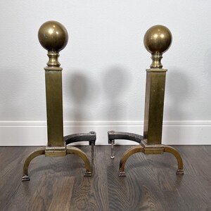 May include: Pair of antique brass and iron fireplace andirons. Each andiron features a brass ball finial atop a rectangular brass column. The base has curved brass legs and a black iron support for holding logs. The andirons are displayed on a dark wood floor.
