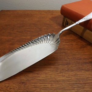 May include: A silver serving utensil with a decorative, scalloped edge and a long, ornate handle. The utensil is resting on a wooden surface.