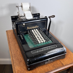 May include: Vintage black Burroughs adding machine with white number keys and a paper roll. The machine has a handle on the side and the Burroughs logo on the front. It sits on a wooden table.