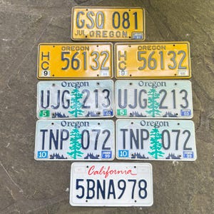 May include: A collection of vintage license plates from Oregon and California. The Oregon plates are yellow, white, and blue, featuring numbers and letters. One California plate is white with red text. The plates are arranged on a gray surface.